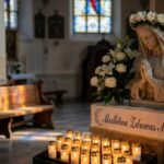 Obraz przedstawiający rzeźbę Matki Boskiej z bukietem kwiatów na głowie i białymi kwiatami wokół niej, a także świecami. Obraz znajduje się wewnątrz pustego kościoła.
