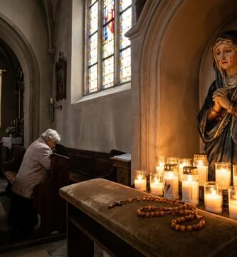 Obraz przedstawiający niewielki postument w kościele, na którym znajduje się popiersie Matki Bożej Bolesnej otoczone świecami i różańcem; w tle widać grupę modlących się starszych ludzi