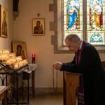 Obraz przedstawiający księdza modlącego się na kolanach w kościele przed zapalonymi świecami i obrazem Jezusa na krzyżu