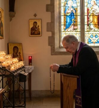 Obraz przedstawiający księdza modlącego się na kolanach w kościele przed zapalonymi świecami i obrazem Jezusa na krzyżu