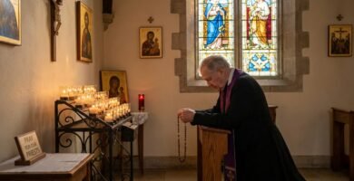Obraz przedstawiający księdza modlącego się na kolanach w kościele przed zapalonymi świecami i obrazem Jezusa na krzyżu