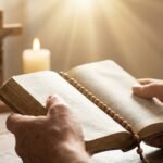 Zbliżenie dłoni osoby trzymającej otwartą Biblię, z której pada światło; w tle stół z krzyżem i świecą