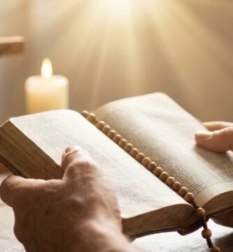 Zbliżenie dłoni osoby trzymającej otwartą Biblię, z której pada światło; w tle stół z krzyżem i świecą