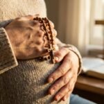 Zbliżenie na brzuch kobiety w ciąży z różańcem w dłoniach, w tle Biblia z krzyżem i świecą