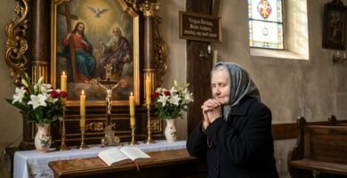 Zdjęcie starszej kobiety modlącej się w kościele obok obrazu Trójcy Świętej, w otoczeniu białych kwiatów, świec i Biblii.