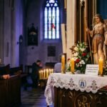 Obraz starszej kobiety modlącej się w kościele, przed nią znajduje się wizerunek archanioła Michała otoczony kwiatami i świecami