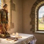 Wnętrze kaplicy z widokiem na stół ze statuą św. Michała Archanioła, świecami i różańcem; w bocznym oknie widać wiejski krajobraz.