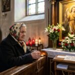 Obraz starszej kobiety modlącej się w kościele, obok niej mały postument z wizerunkiem archaniołów, otoczony białymi kwiatami i świecami